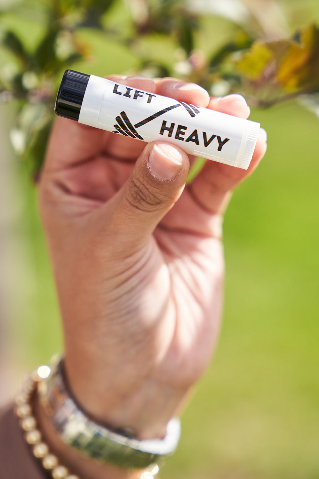 Natural lip balm with lift heavy wording and dumbell on the label. Held in a hand with a blurred tree in the background.