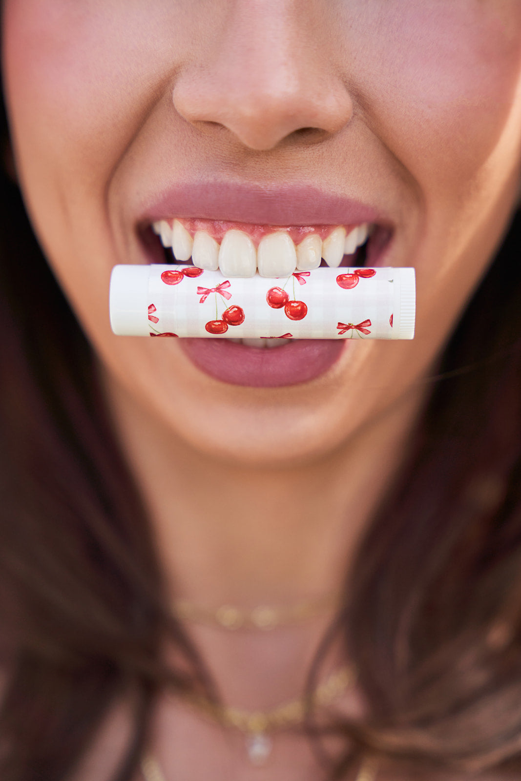 A facial close up. Model holding cherry patterned lip balm between her teeth.