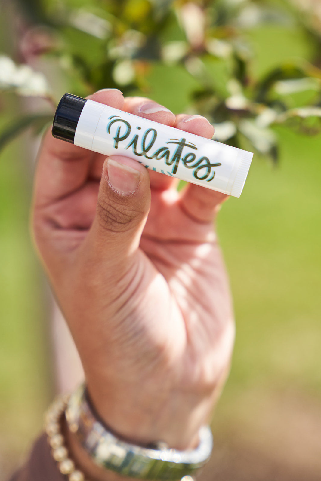 Pilates lip balm. Inhale, exhale. Woman holding it with gold bracelets with a blurred tree background.