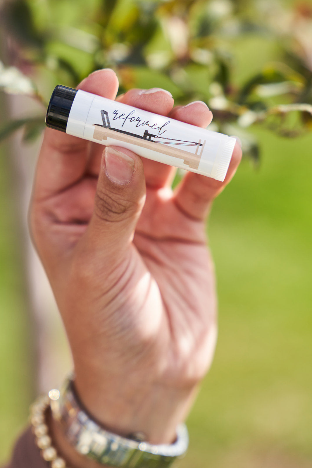 A pilates reformer design on a lip balm that says reformed. Being held in a womans had with gold bracelets. Blurred tree in the background.