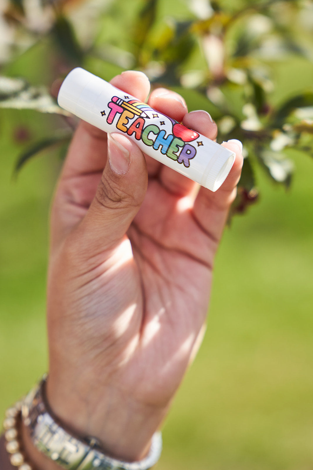 Hand holding a lip balm with 'Teacher' text against a blurred green background