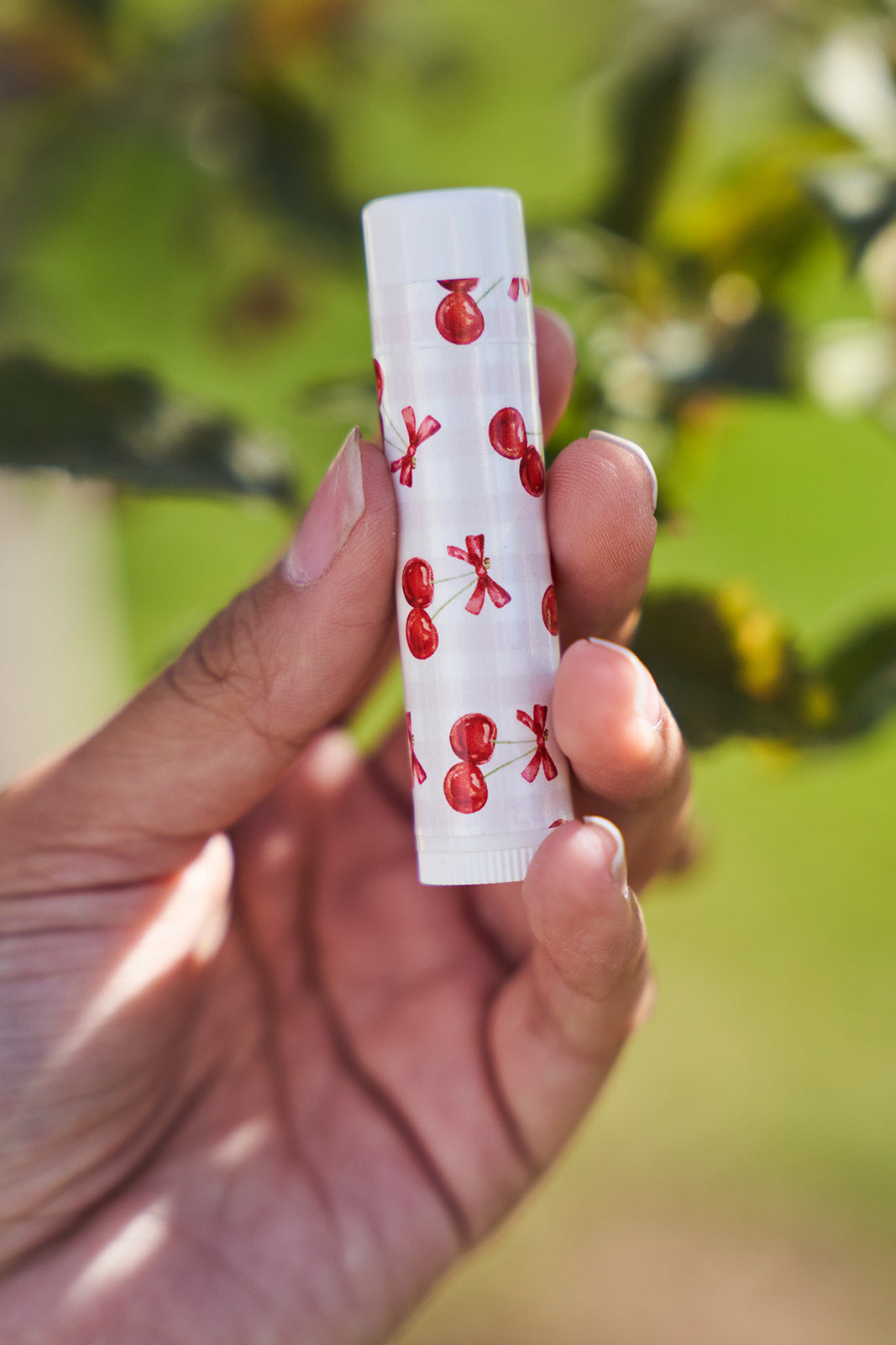 A hand with gold bracelets holding a light pink checkered background with bold cherry lip balm label on a white tube. 