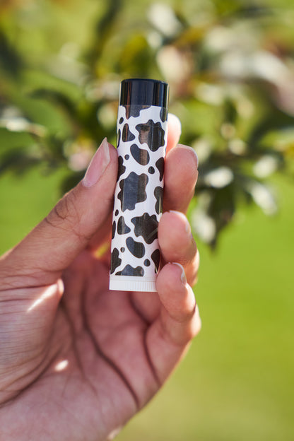 Hand hold a black and white cow print lip balm with a blurred tree in the background. 