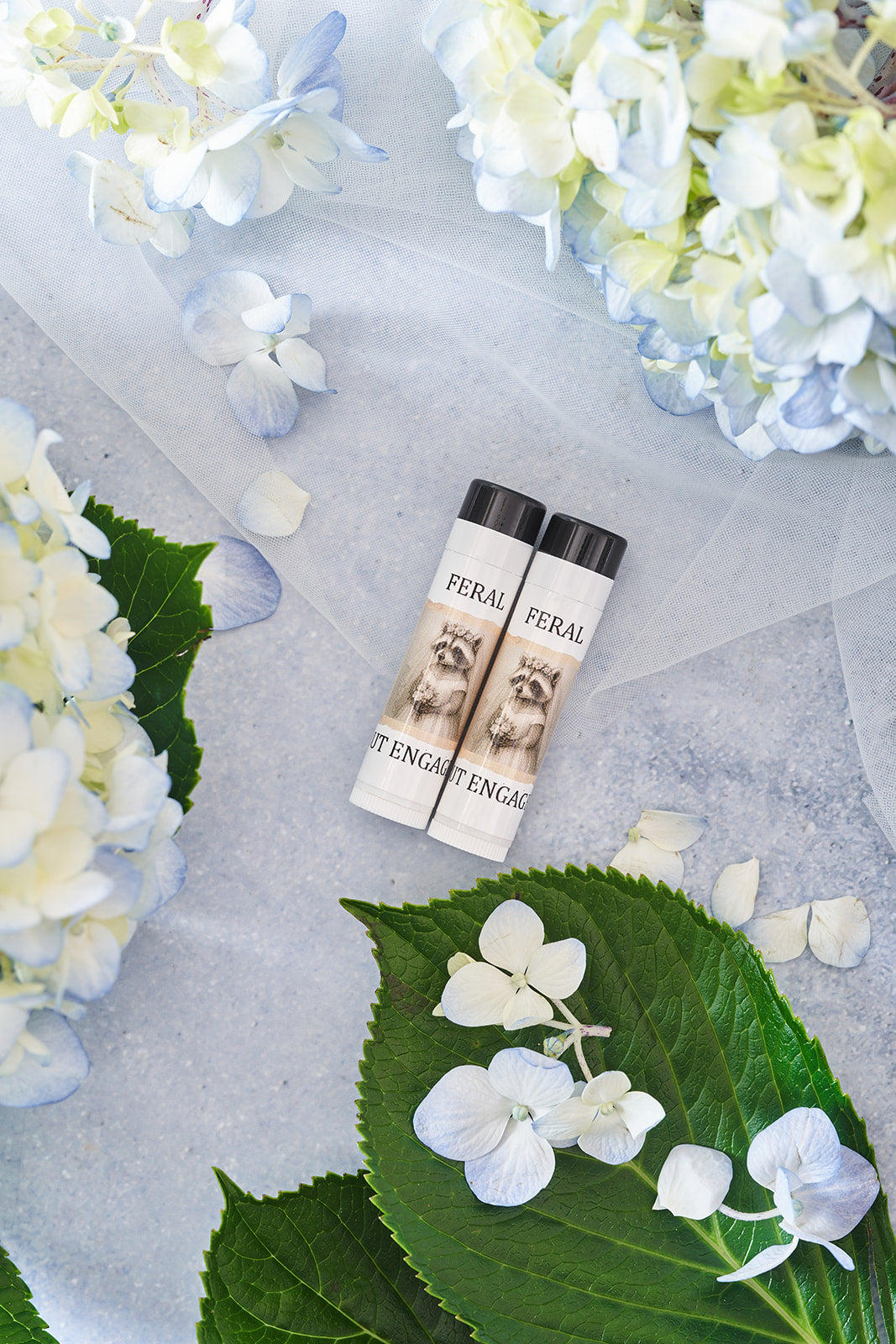 Two lip balms with 'FERAL but engaged' branding on a gray surface with hydrangea flowers.