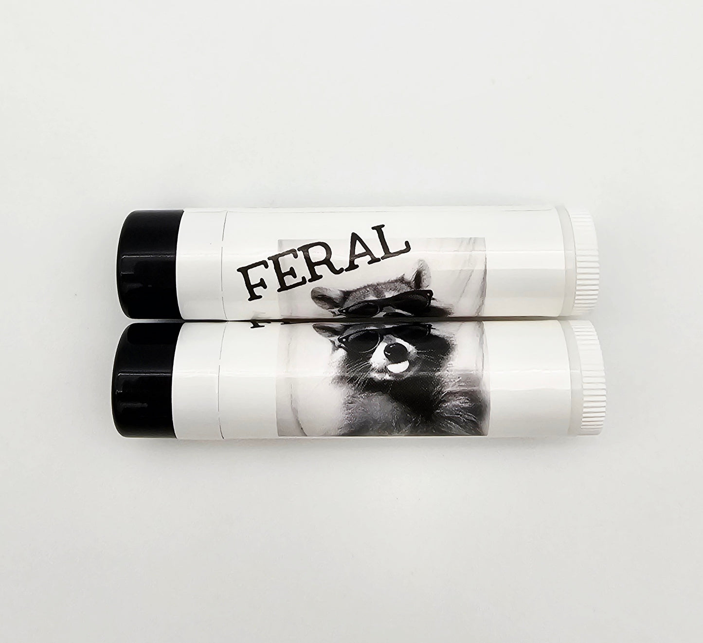 Two lip balm tubes with 'FERAL' branding on a white background. Raccoon wearing glasses sticking out their tongue 