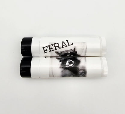 Two lip balm tubes with 'FERAL' branding on a white background. Raccoon wearing glasses sticking out their tongue 