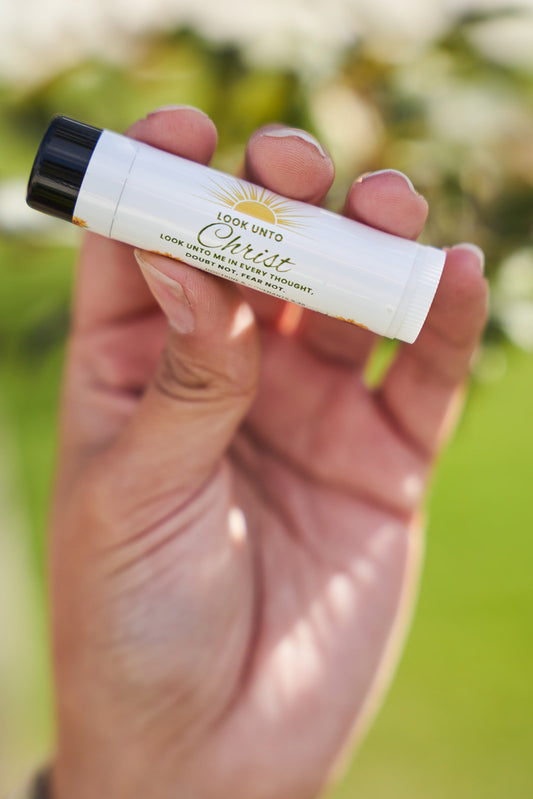 Hand holding a small white lip balm tube that says look unto christ. Yellow sun and sunflower design with black cap against a blurred green background