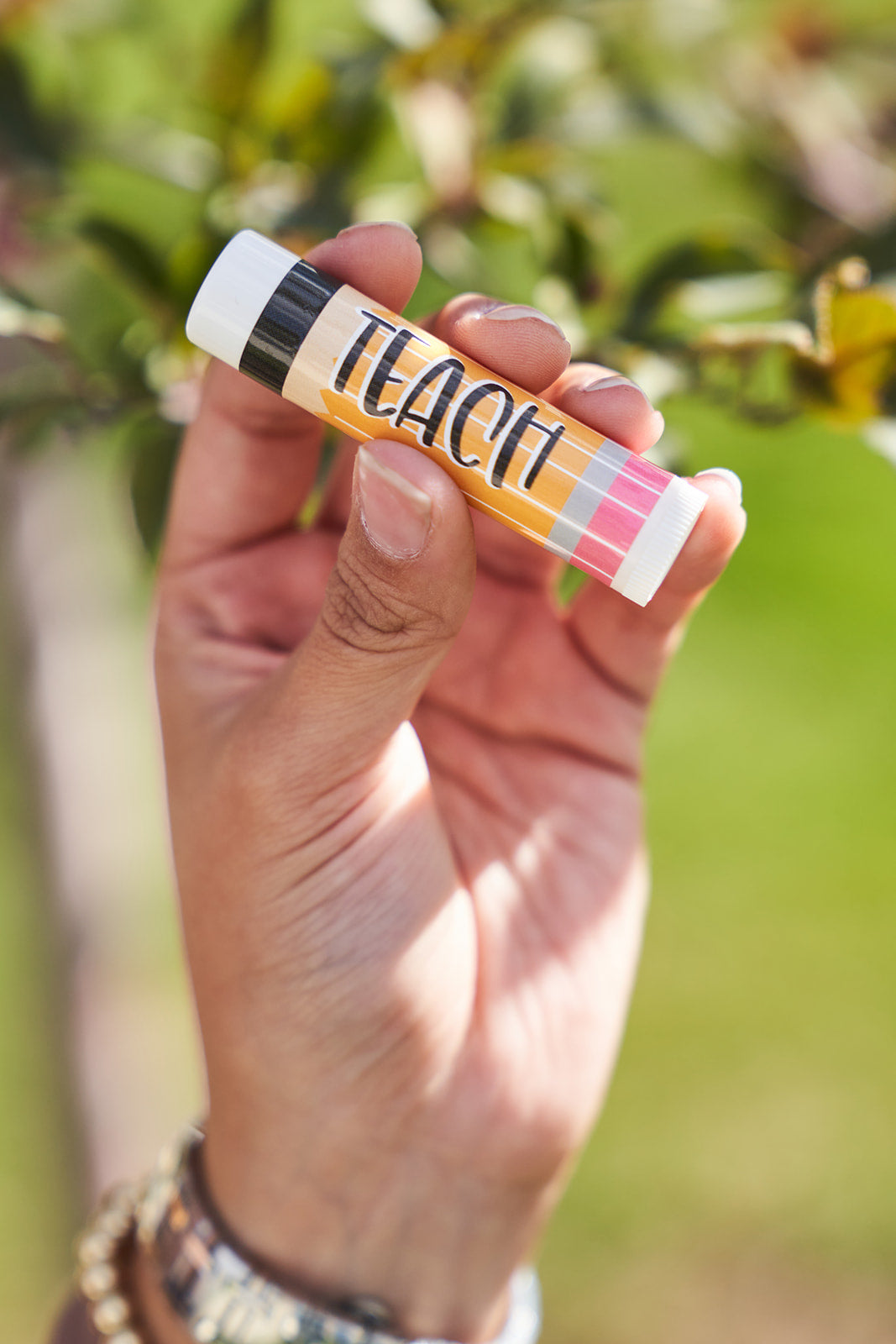 Hand holding a colorful lip balm with 'TEACH' written on it looking like a pencil against a blurred green background
