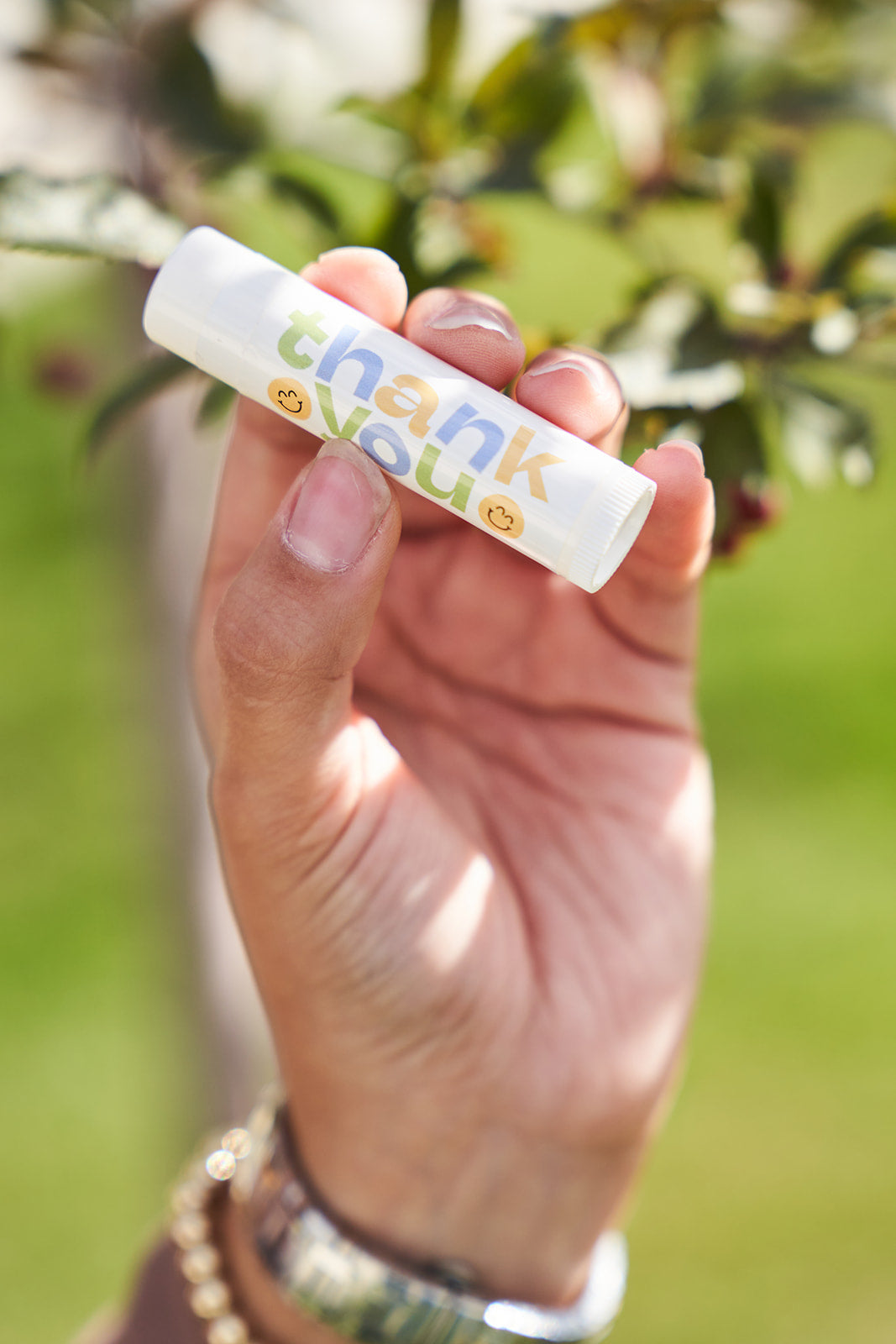 Thank you lip balm with green yellow and blue with smily face on a white background. A hand with gold bracelets holding it. 